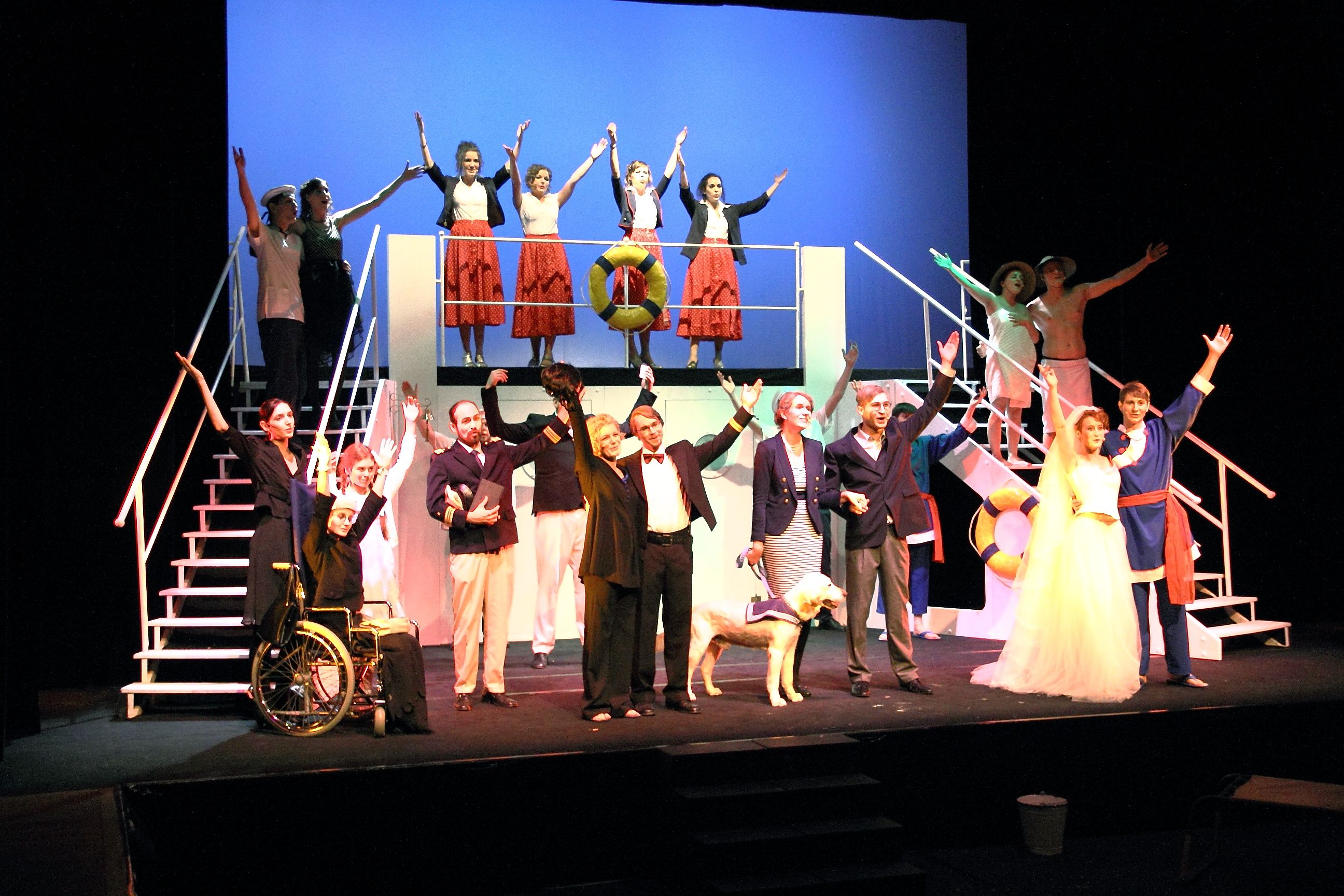 Ein Gruppenfoto einer Musical-Aufführung: alle Schauspieler stehen auf der Bühne und heben ihre Hände in der Luft. Auch ein Hund ist dabei.